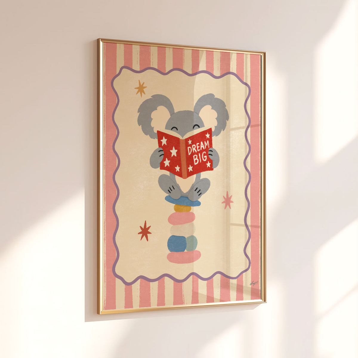 Illustrated grey koala sitting on stacked stones reading a red Dream Big book, inside a wavy border on a pink and cream striped background.