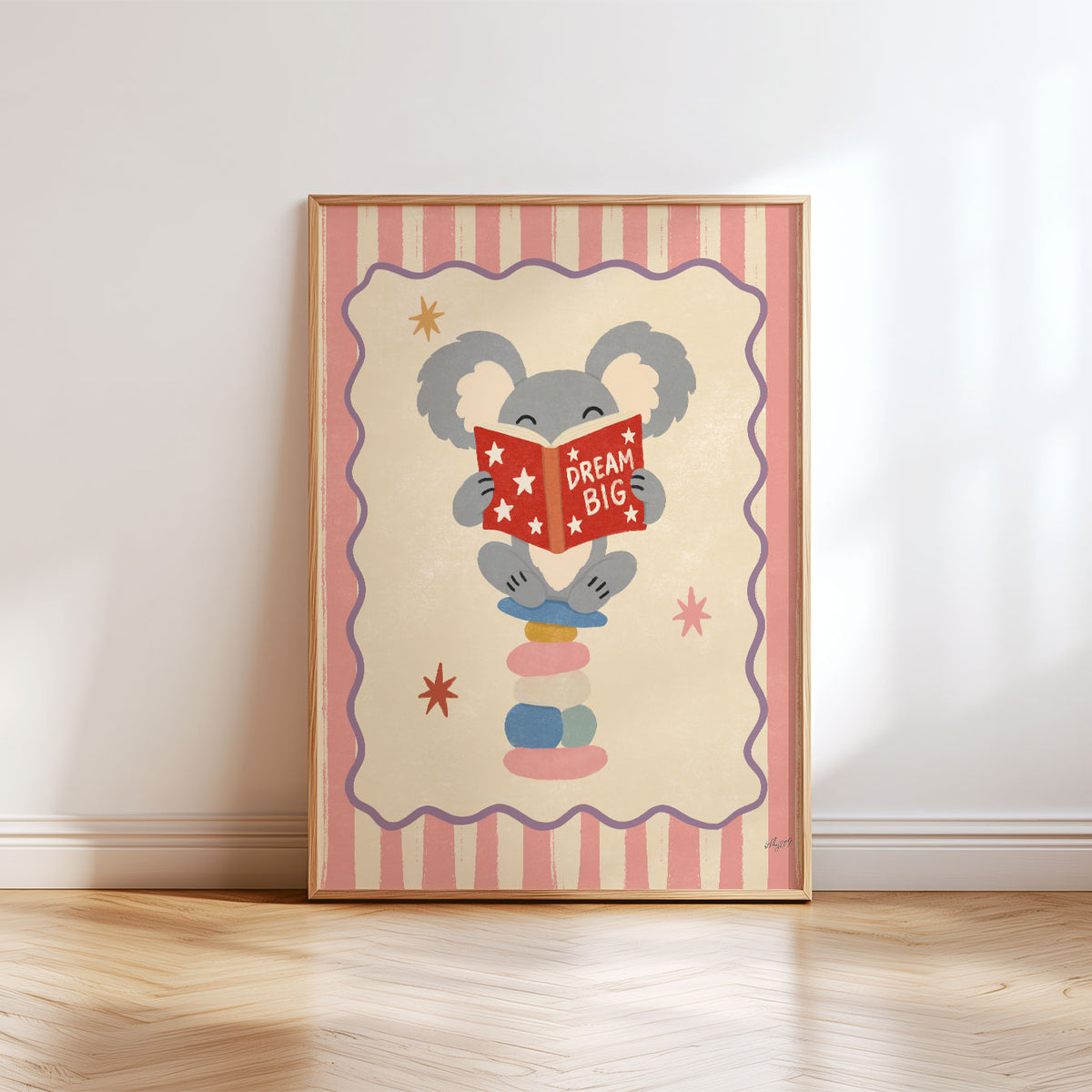 Illustrated grey koala sitting on stacked stones reading a red Dream Big book, inside a wavy border on a pink and cream striped background.