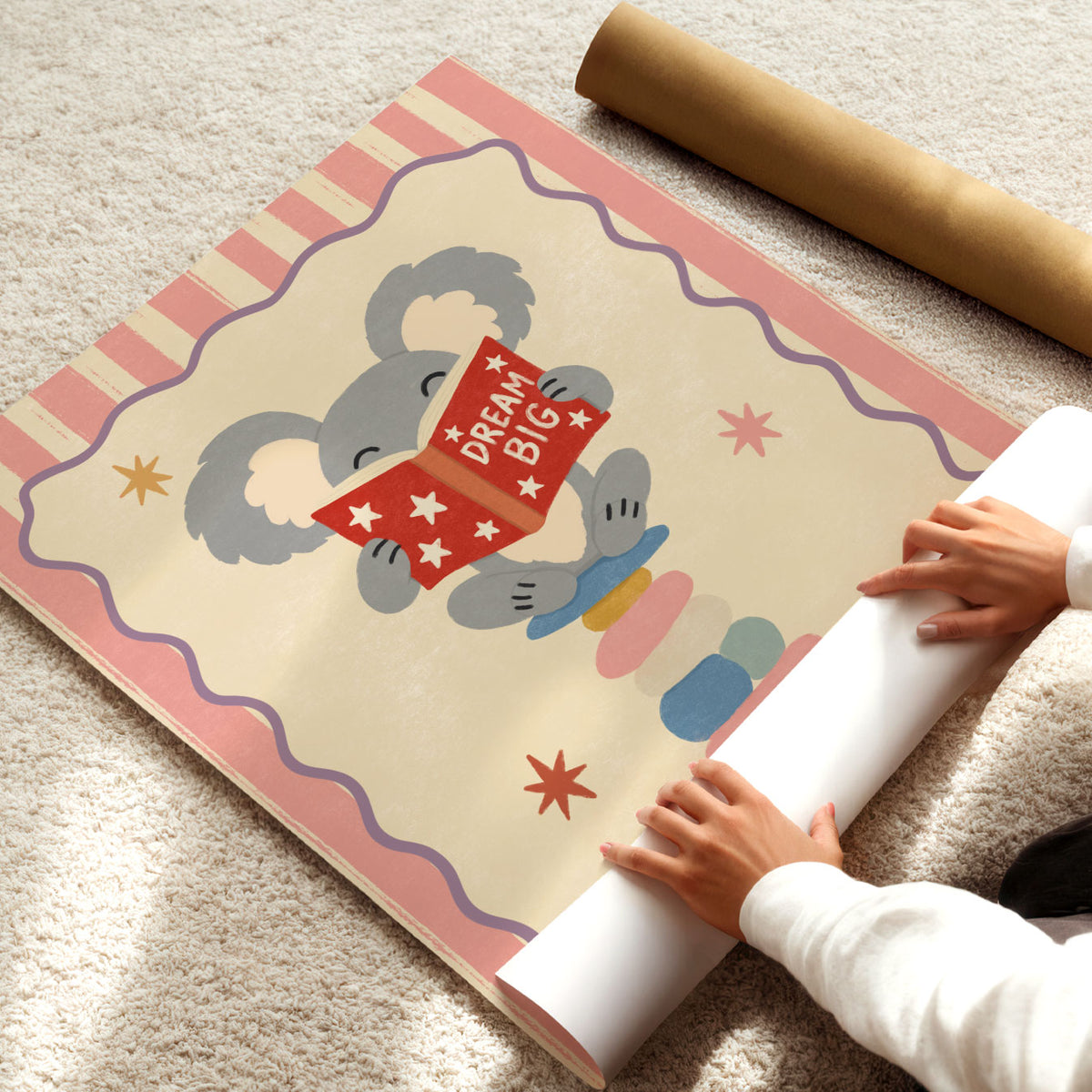 Illustrated grey koala sitting on stacked stones reading a red Dream Big book, inside a wavy border on a pink and cream striped background.