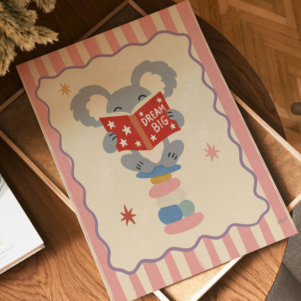 Illustrated grey koala sitting on stacked stones reading a red Dream Big book, inside a wavy border on a pink and cream striped background.